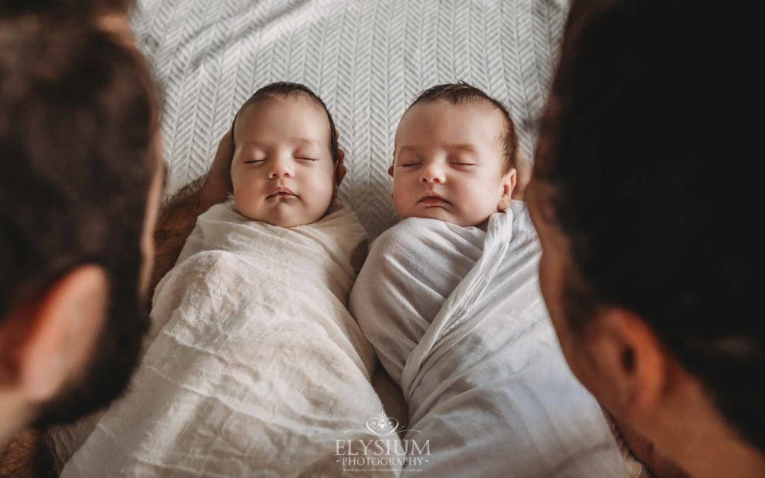 Parents hold their newborn twins between them as they lay on a patterned white blanket