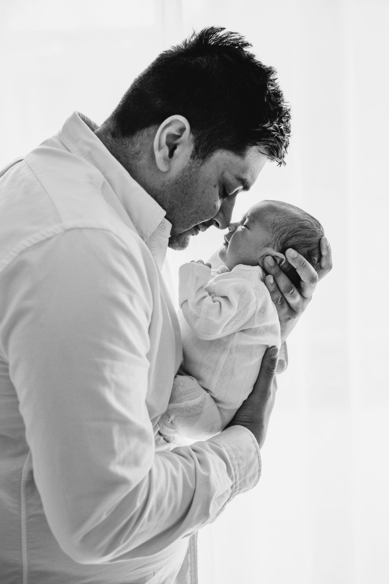A father presses his nose gently against his newborn baby's face in front of a bright window