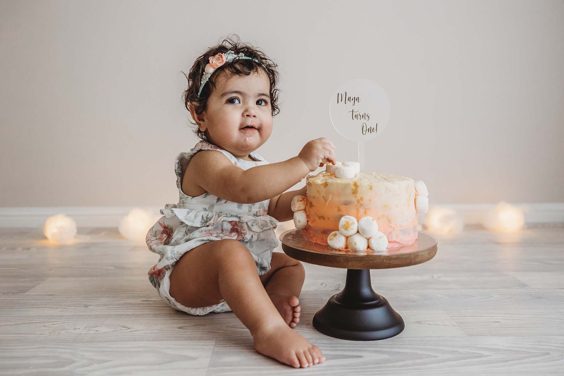 A baby girl sits with her birthday cake before smashing it wearing a floral romper