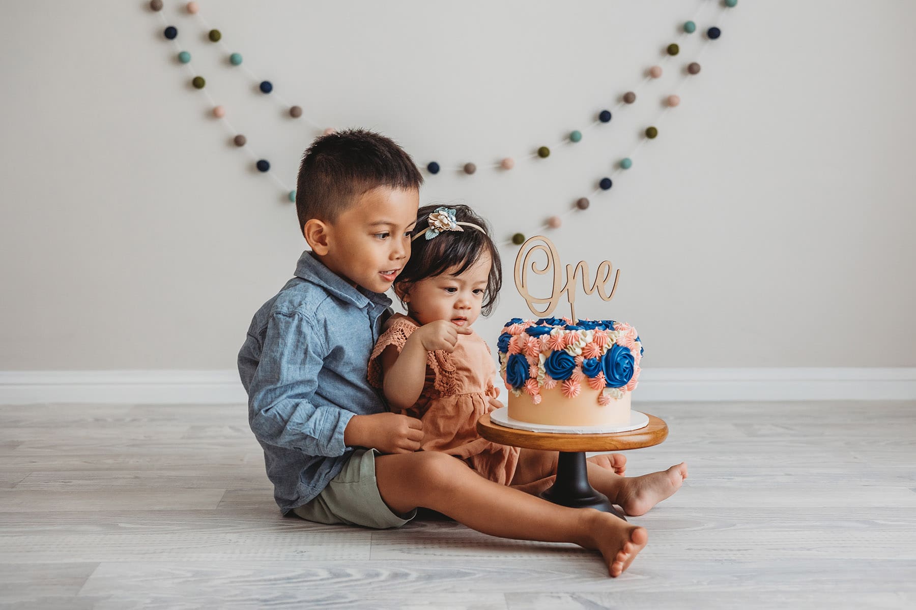 A baby girl sits with her big brother in front of a colourful cake she's about to smash for her first birthday