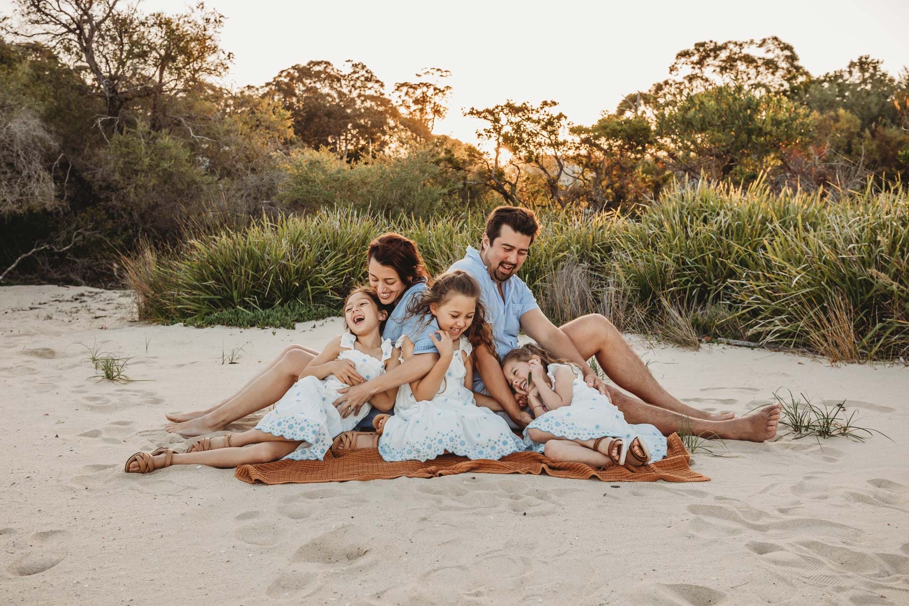 Parents sit on a blanket tickling their 3 girls as the sun sets over the beach