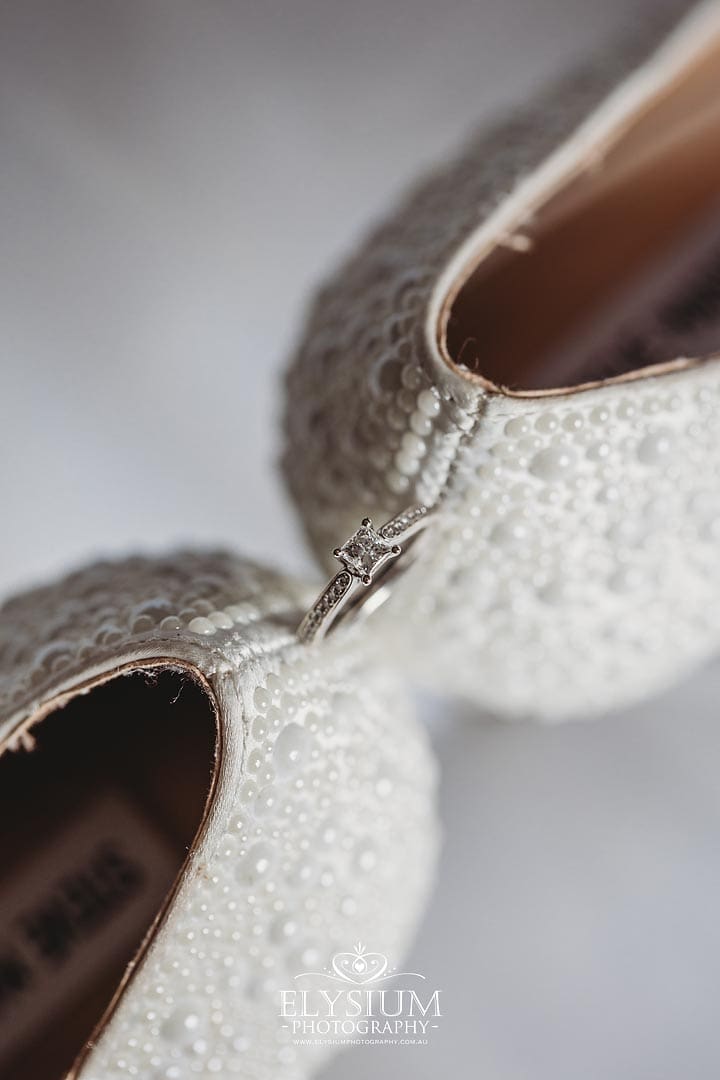 A brides engagement ring sits balanced on the heel of her wedding shoe