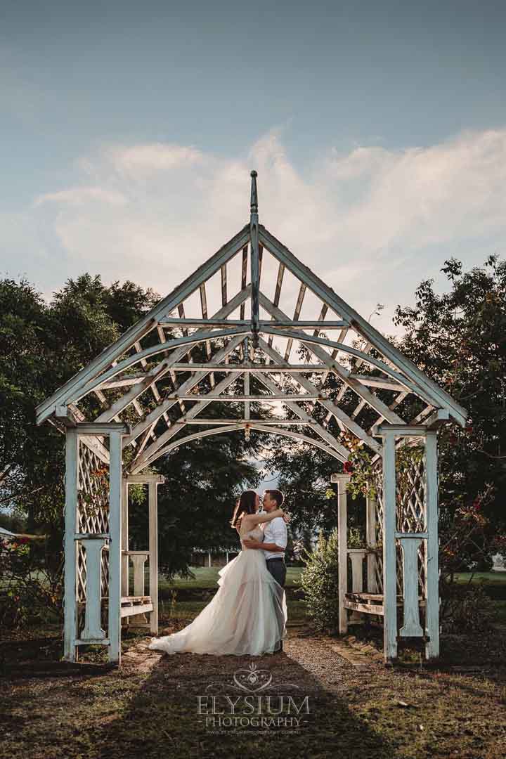 Newlyweds stand hugging under a white arbor backlit by a flash at sunset 