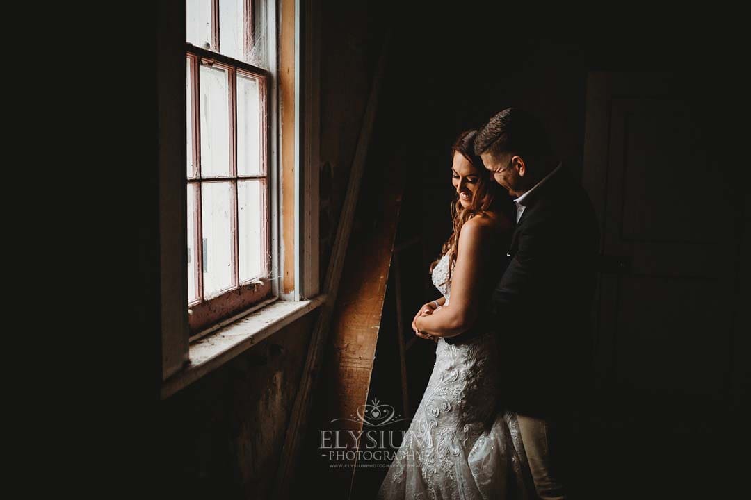 A couple smile as they cuddle next to an old window in a darkened room of a homestead