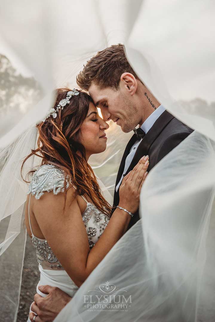 A married couple stand under the brides veil and hug as the sun sets behind them