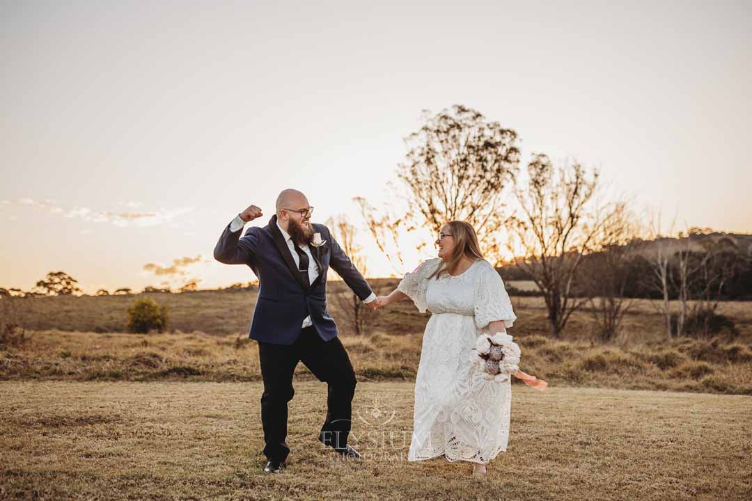 A bride and groom stand cheering in a grassy field at Ottimo house as the sun sets behind them