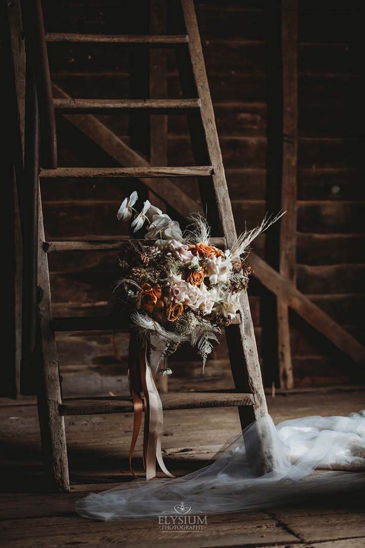 A bridal bouquet with muted orange tones sits on a rustic ladder in a barn 