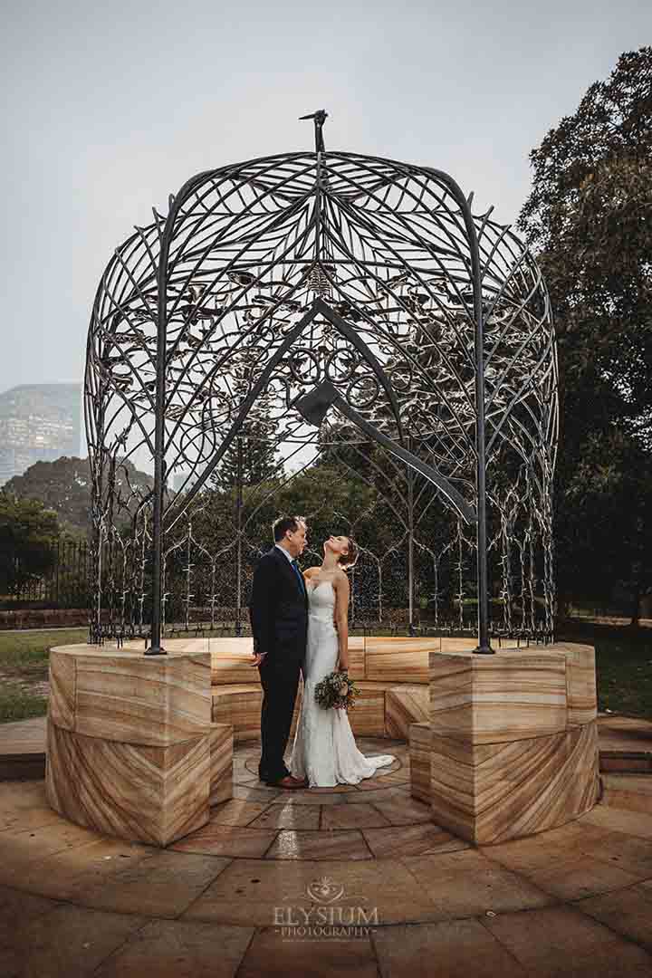 A married couple stand hugging under an iron rotunda as rain begins to fall on them