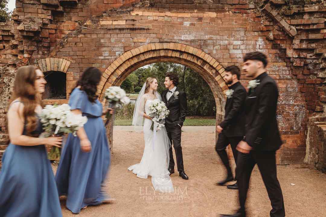 Newlyweds stand next to a brick arch as their bridal party walk around them with motion blur