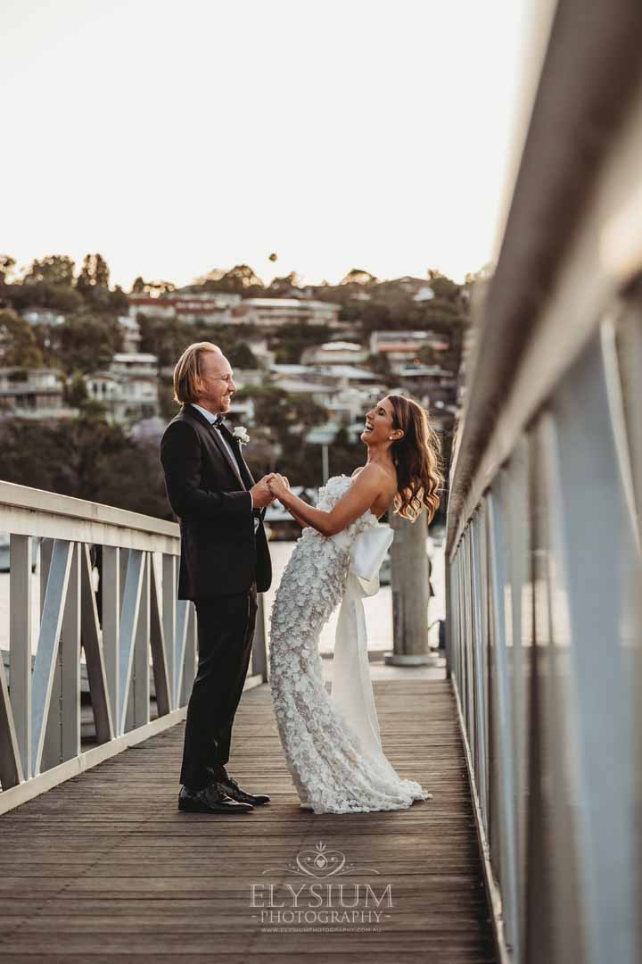 A bride and groom stand on a pier and laugh as the sun sets behind them over Sydney harbour