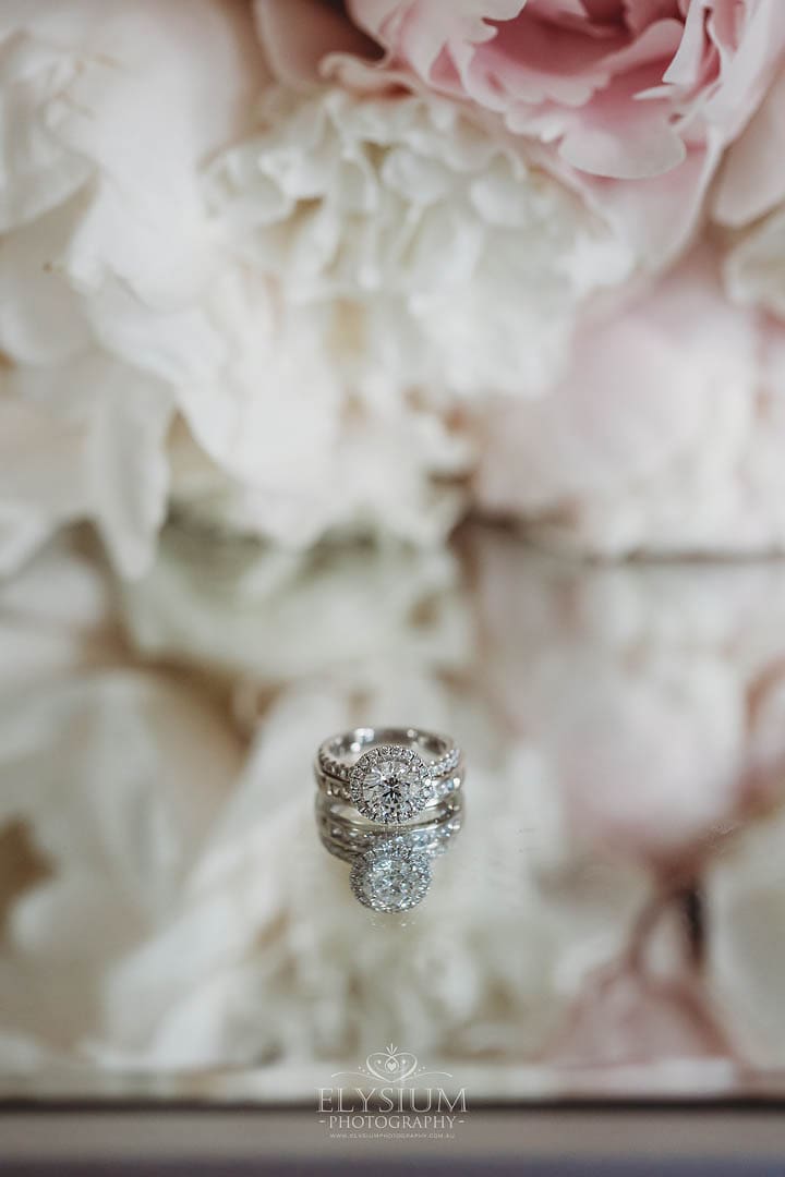 A brides engagement ring sits on a mirrored table with the bouquet blurred in the background