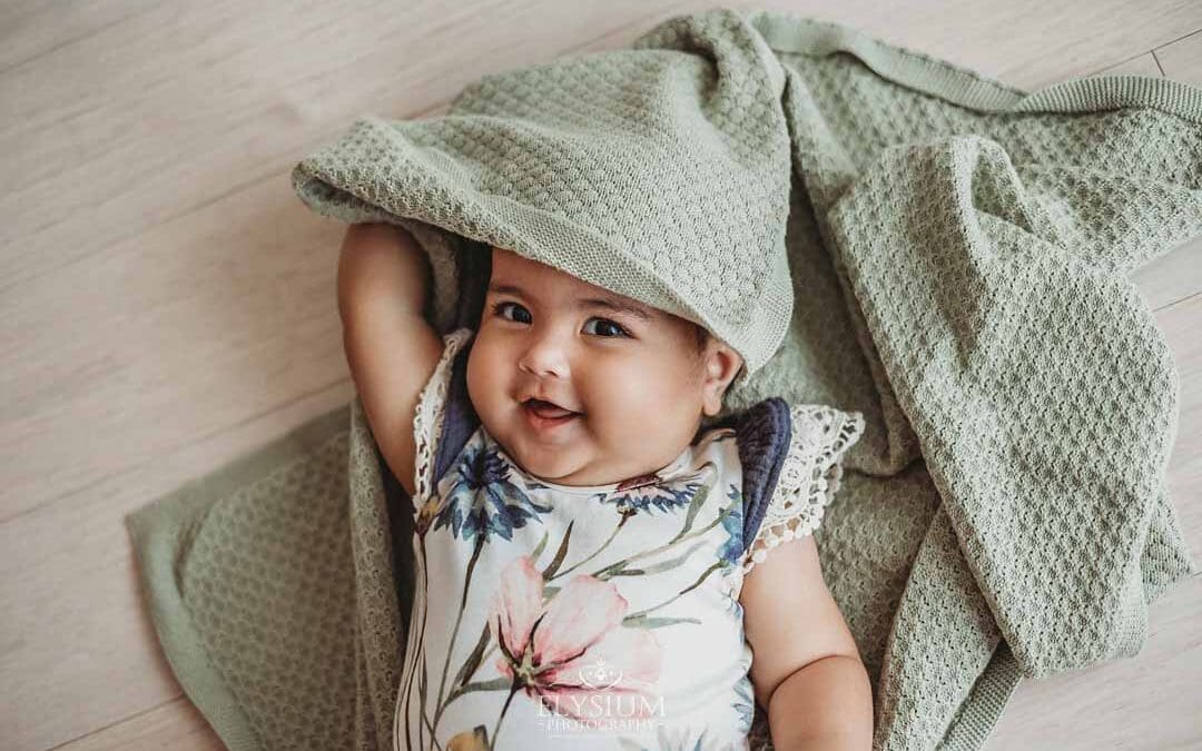 A baby girl dressed in a floral romper lays on her back and smiles peeking out from under a green blanket