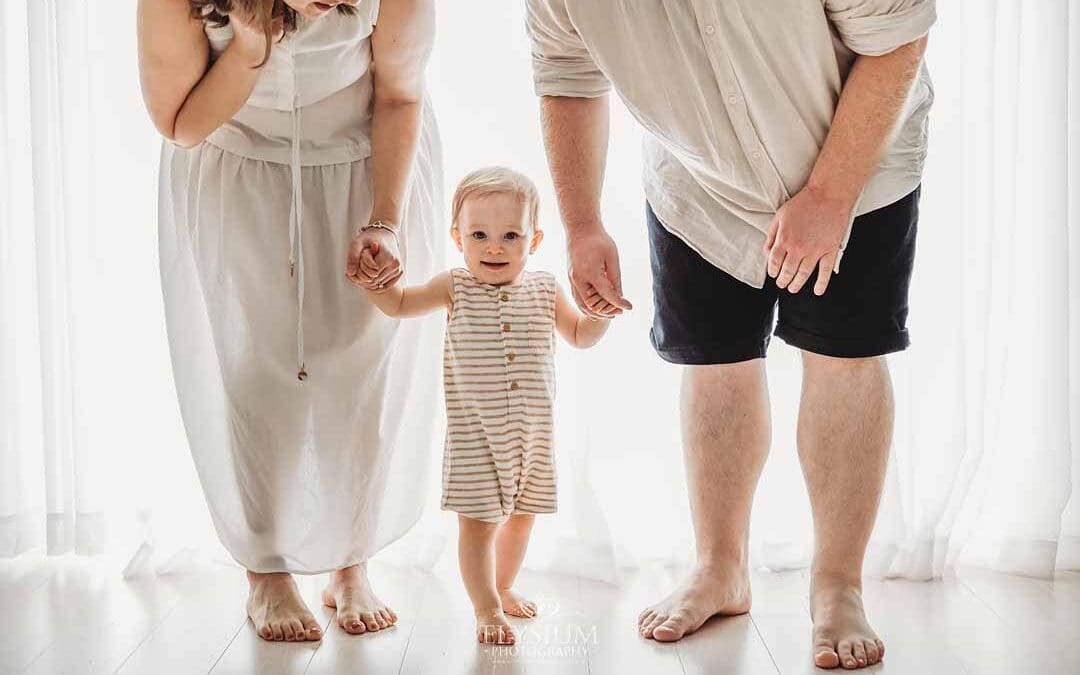 A baby boy stands between his parents holding their hands in front of a large studio window