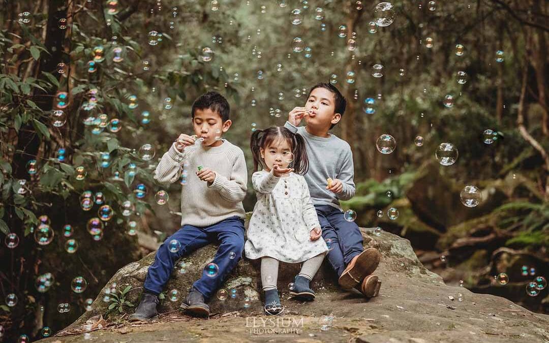 Three siblings sit on a huge rock surrounded by trees and blow bubbles